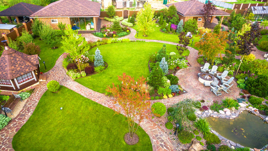 An aerial view of a professionally landscaped backyard featuring winding stone walkways, a circular brick patio with a fire pit, a small pond, and various ornamental trees.