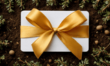 A white gift card wrapped in a large gold satin bow, resting on dark brown soil framed by small green sprigs.